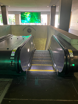 A picture of an escalator entrance. In the foreground there is an escalator and in the background slightly to the left of center for the image there is a large video board that is green. Between the video board and the top of the escalator there is a wall that surrounds the escalator bay. There are also three pillars present holding up the ceiling around the wall.